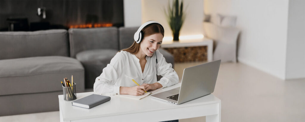 A woman wearing headphones looking at a computer screen and taking notes in a well-light background