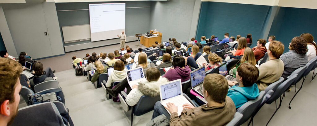 A professor in front of a slide, standing at the bottom of a lecture hall filled with lines of students