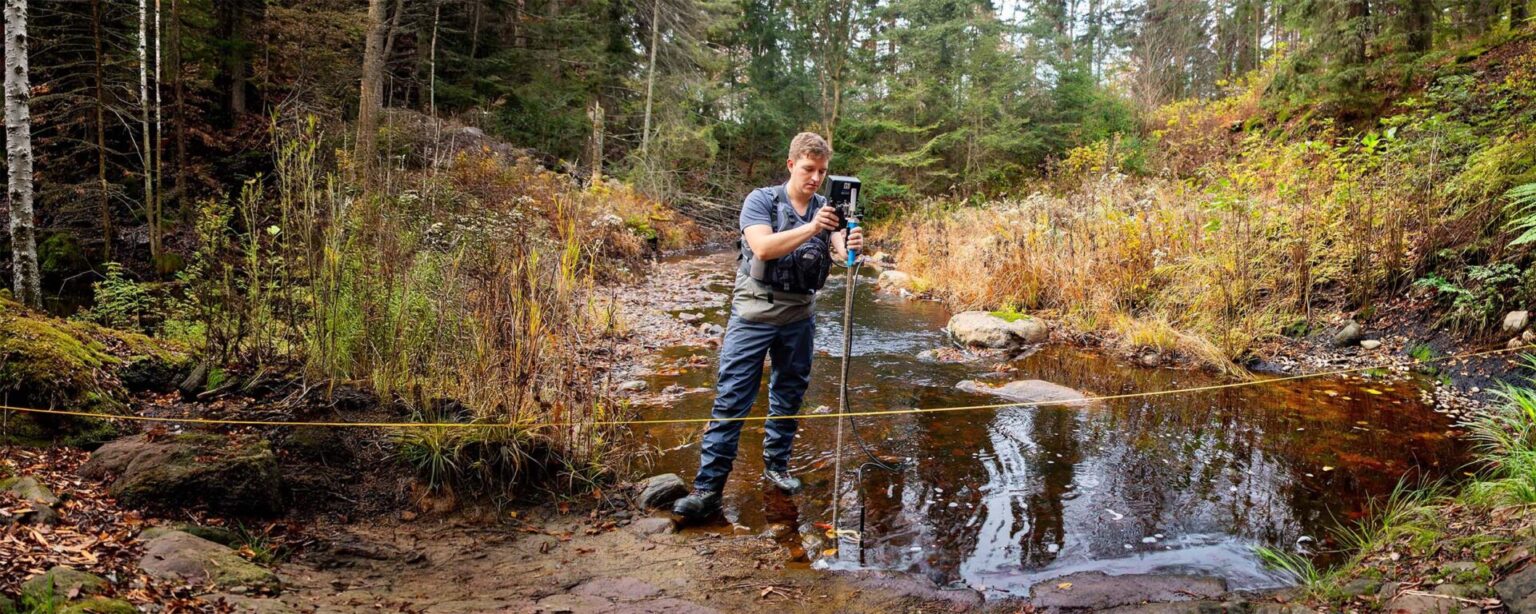 A student standing near the water in nature, measuring something on a device