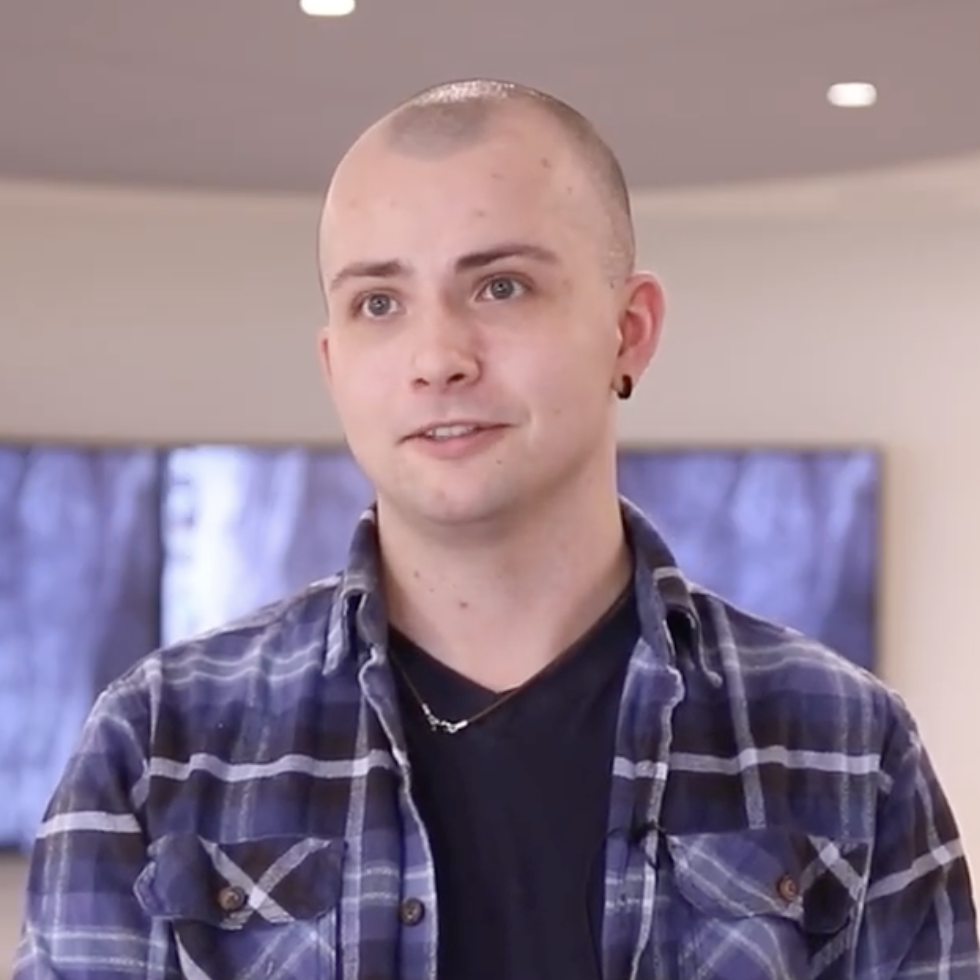 A man in blue plaid shirt in front of a bright beige background with screens