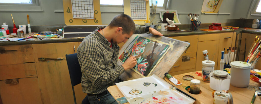A male student painting on a canvas with art supplies in front of him