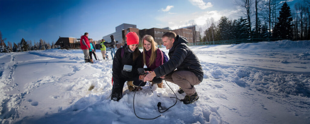 A professor gathered with 2 students looking at a device in the winter with the campus building in the background