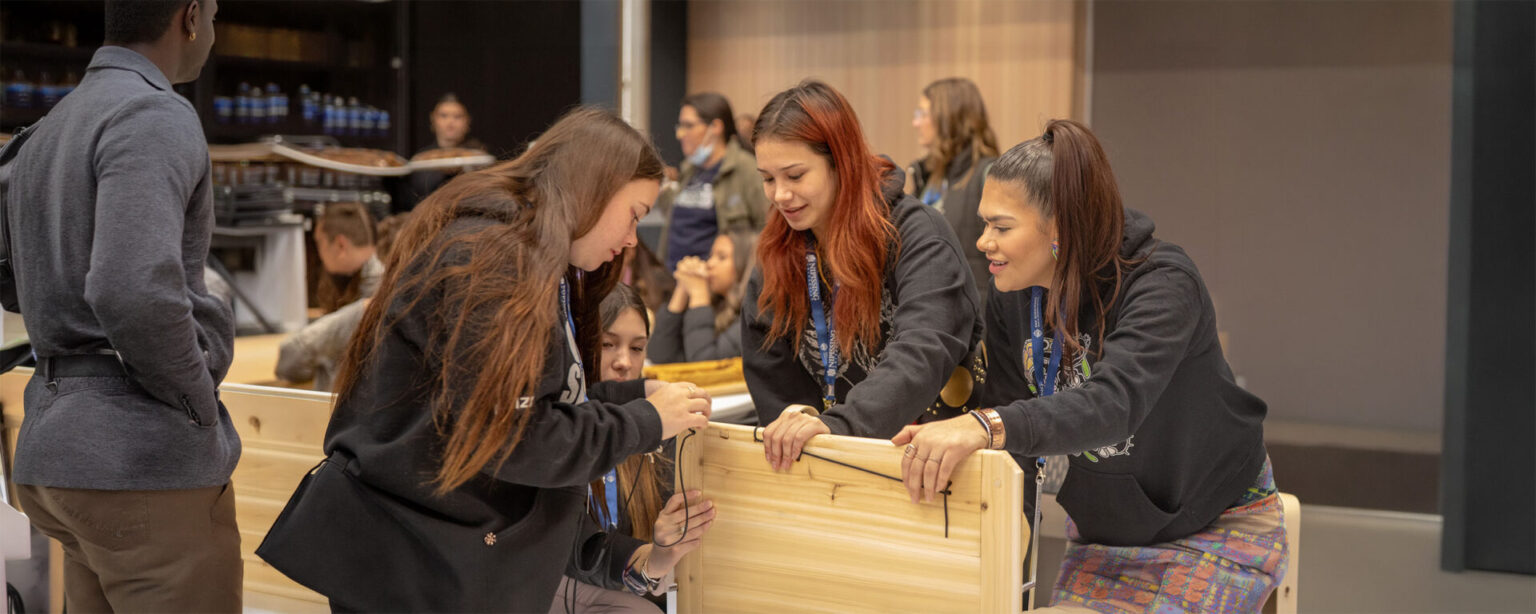 4 students gathered to build something out of wood with many people in the background