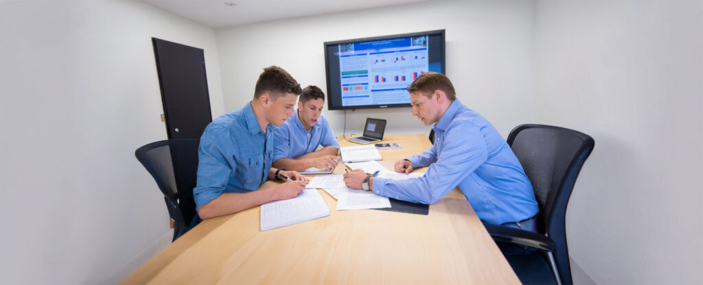 2 students and a professor in blue shirt working together at a table with a monitor screen showing charts behind them