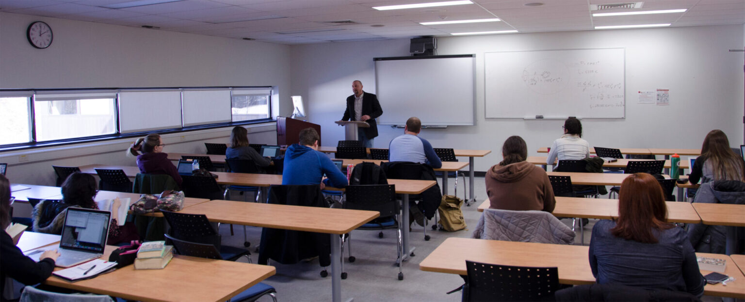 A classroom of students facing the front with a professor doing a lecture
