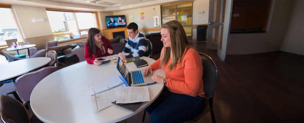3 students working on their laptop on a table in a cafeteria setting