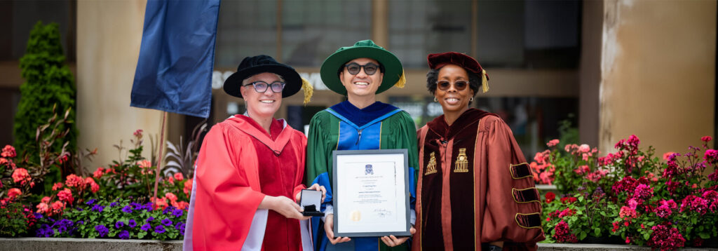 A graduate in green gown holding a plaque with 2 faculties standing on either of his side