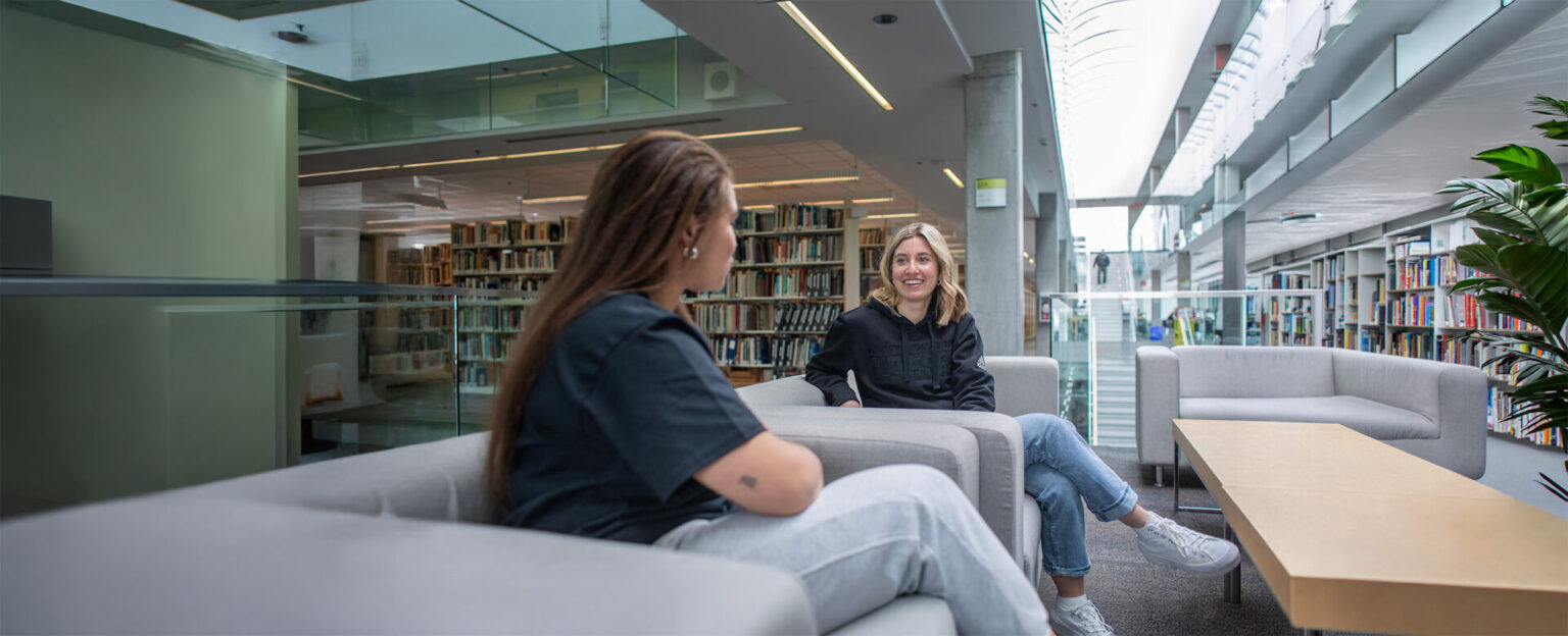 2 students talking, seated in the library couch with the book shelves and library stairs in the background