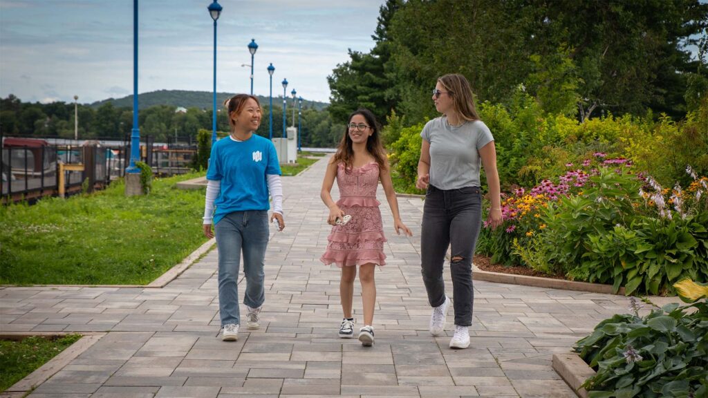 3 female students walking down the stone paved walkway with the waterfront in the background