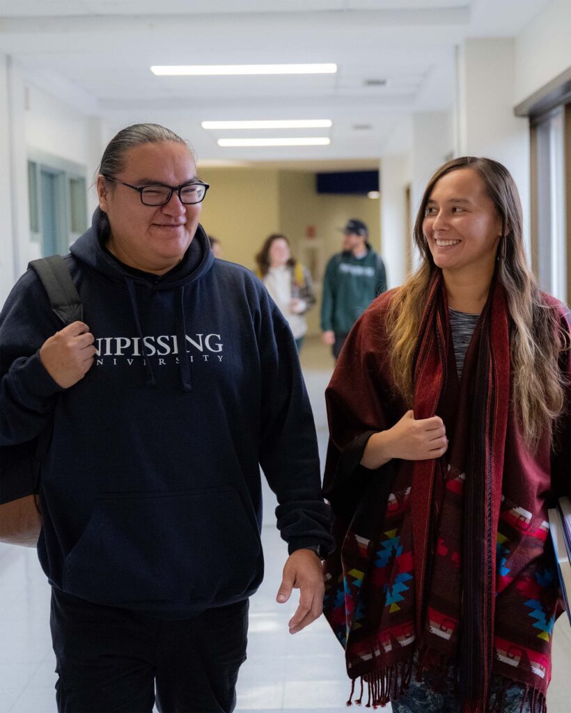 A male student in navy hoodie and a female student in traditional indigenous clothing walking through the hallway
