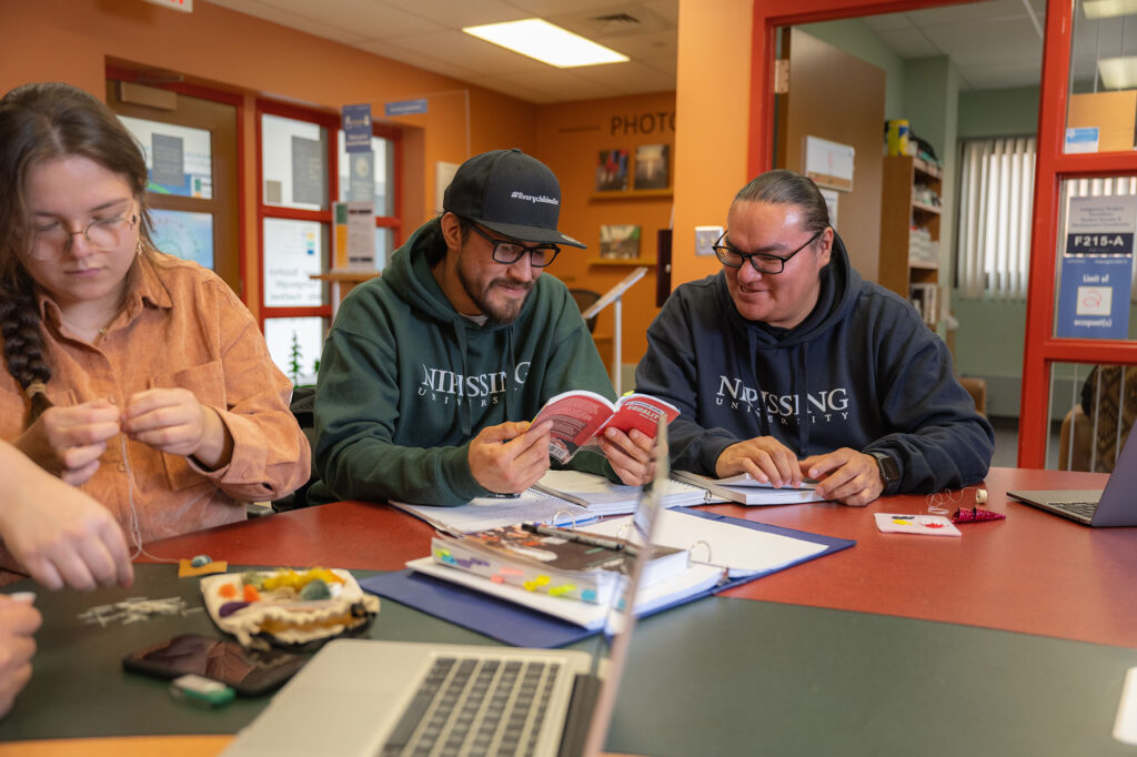 2 male Indigenous students looking at a book while a female student is doing something on their left in the Indigenous office