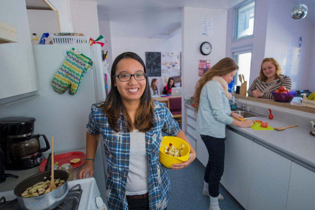 A female student in plaid shirt cooking on the stove while some female students are cutting vegetable in the dorm