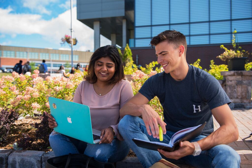 2 students studying on their laptop with a bush and the library in the background