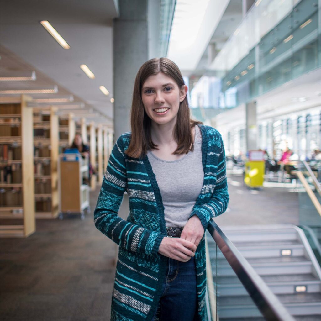 A woman in grey and stripped blue cardigan leaning against a staircase in the library background