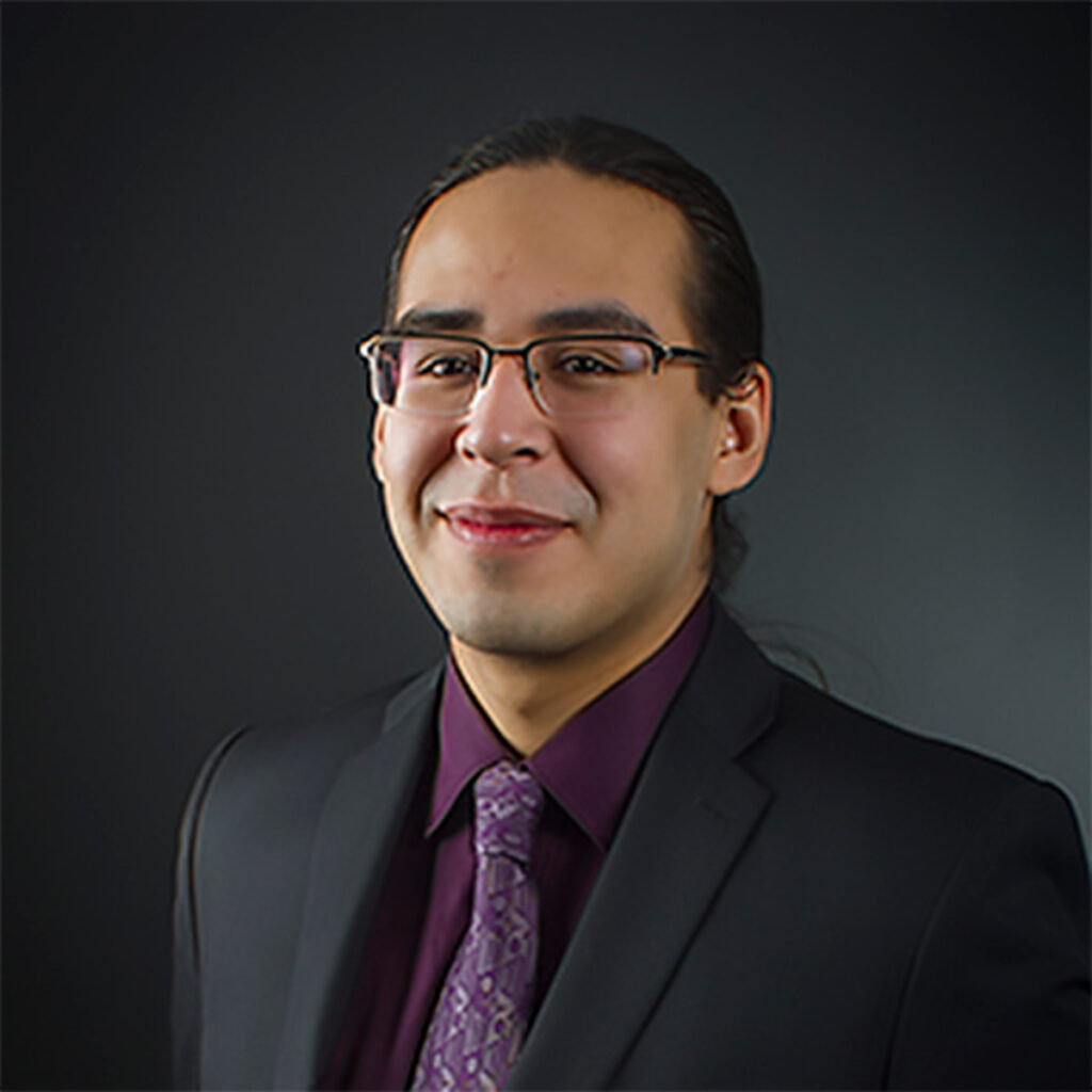 A man with glasses wearing a purple and dark grey suit against a dark background