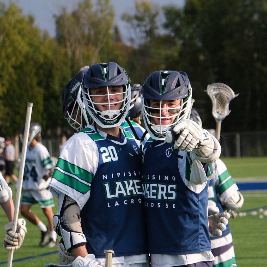 2 students in lacrosse gear point at the camera