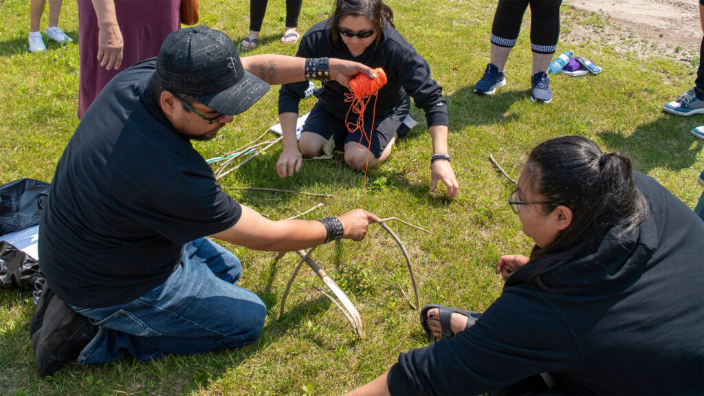 An instructor helping 2 students build something our of twigs on grass