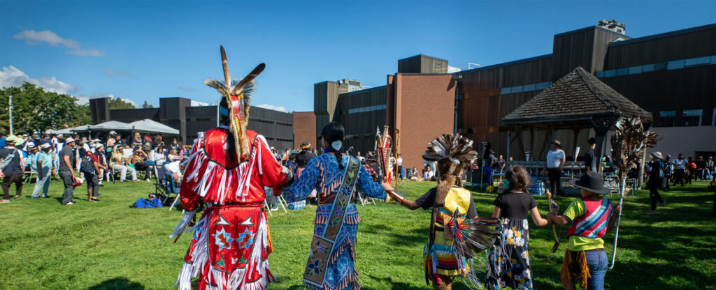 4 people in traditional Indigenous clothing facing the pow wow celebration outdoor