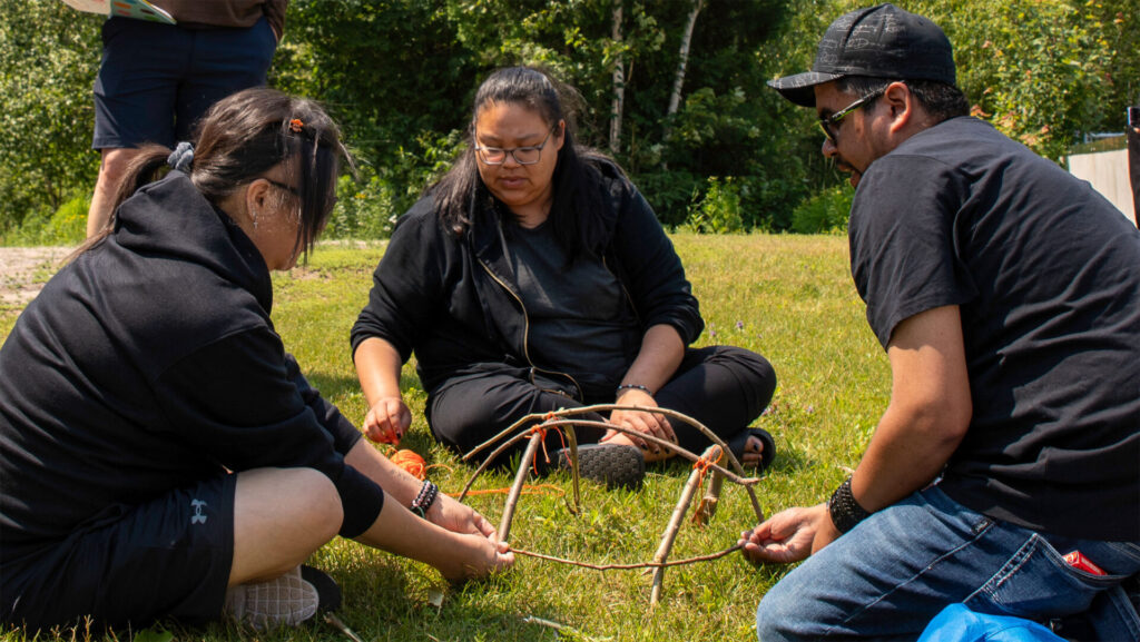An instructor and 2 students sitting on grass building something of twigs