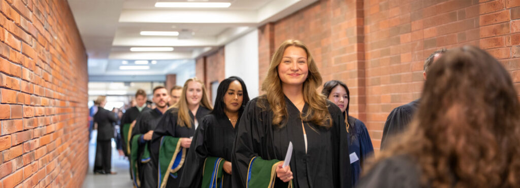 A female student in graduation gown with a line of other graduates behind her walking down the hall