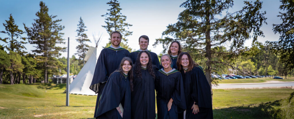 A group of friends in convocation gowns with the tipi in the background
