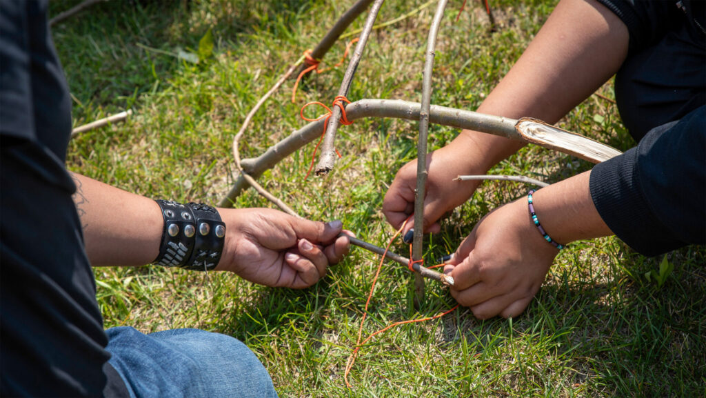 A close up of the arms of people participating in building something out of twigs on the grass