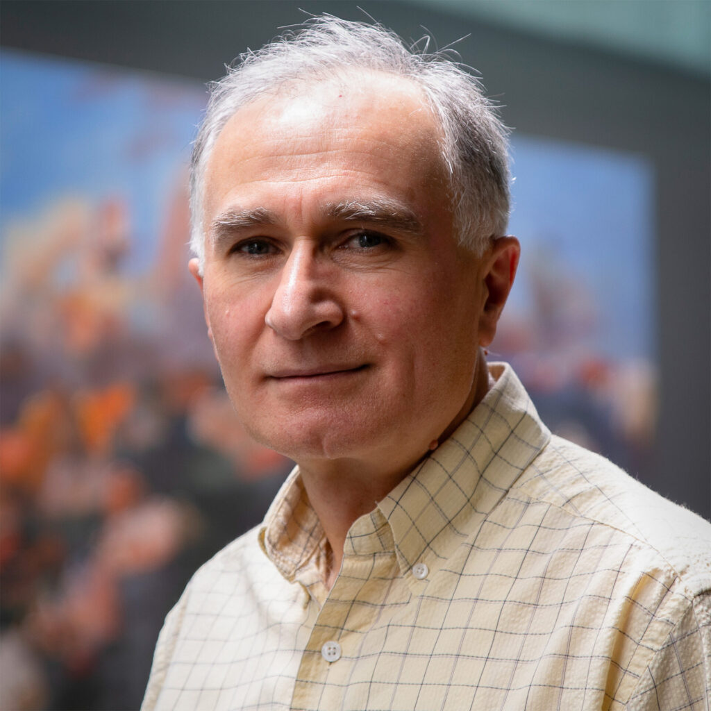 A professor in beige checkered button up standing in front of a painting background