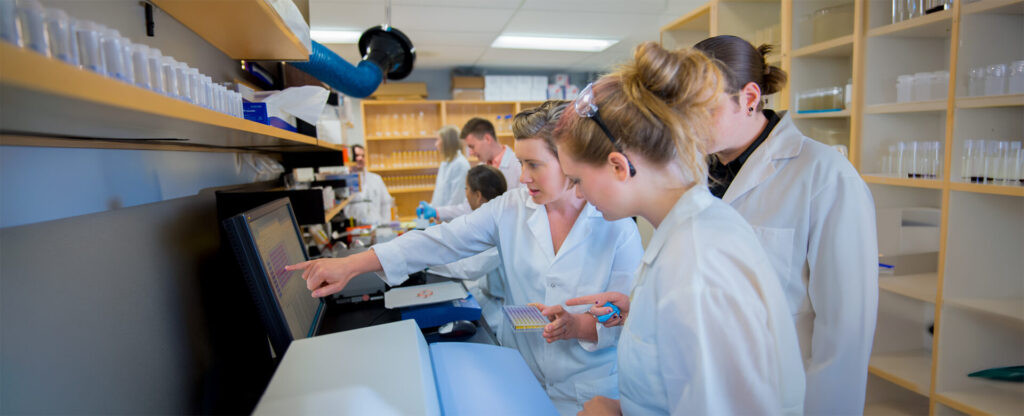 2 students and a professor in white lab coat looking at a monitor screen in the lab environment